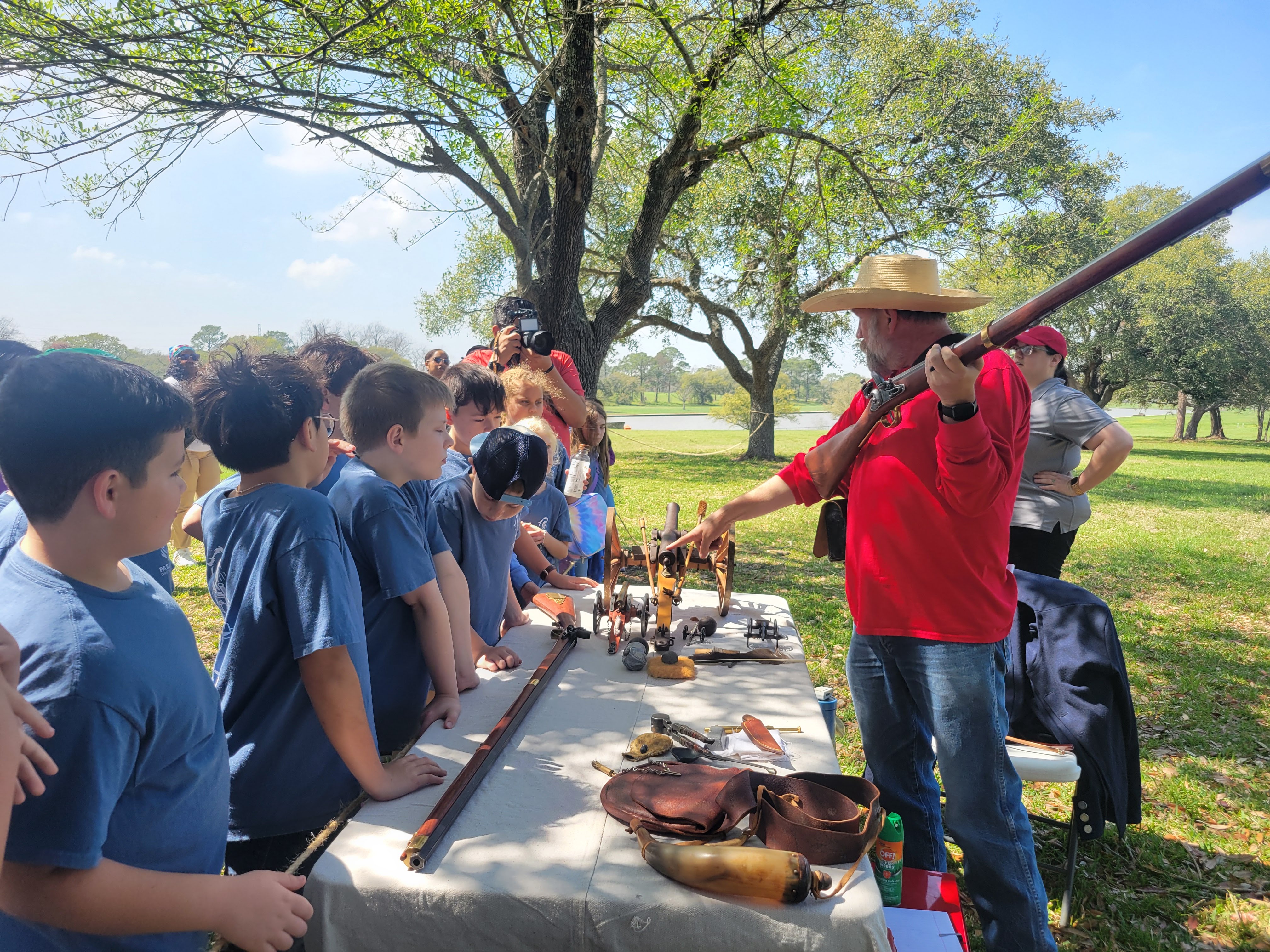 A man holding a reproduction long gun gestures toward a table holding 19th century replicas, while talking to children on the other side of the table. at