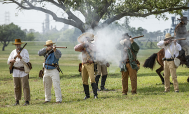 Smoke billows from a line of reenactors firing muskets.