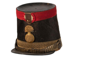 A tall military hat with a red ribbon, a gold medallion, and a gold strap.