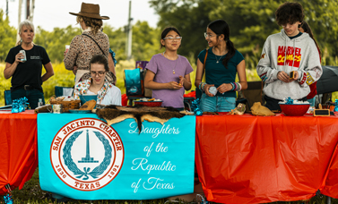 Six people stand behind tables draped with a Daughters of the Republic of Texas banner.