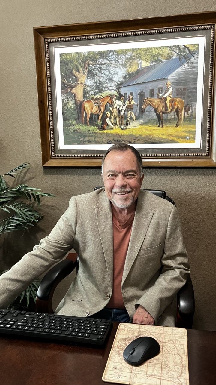 A man seated at a desk with a paiting behind him smiles at the camera.