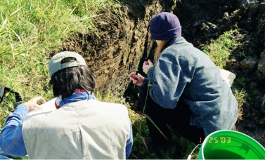 Two archeologists stoop by a trench in the ground.