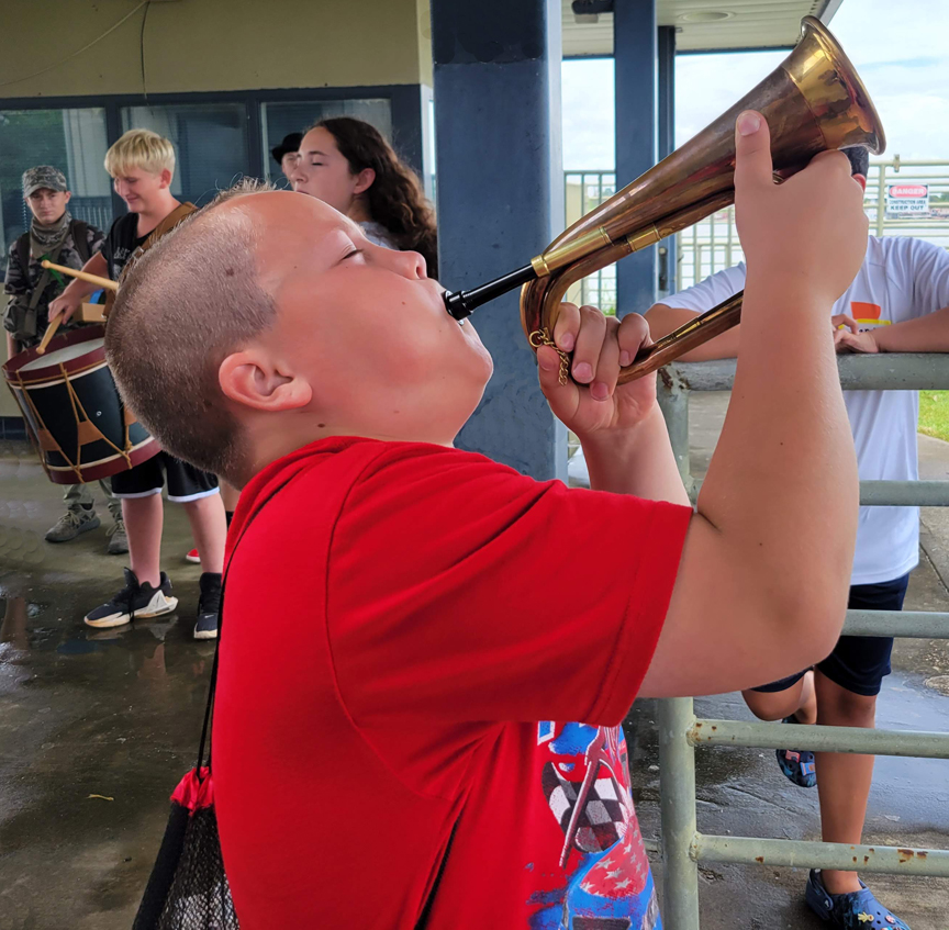 A boy wearing a t-shirt plays a bugle, with a  drum player and spectators in the background.