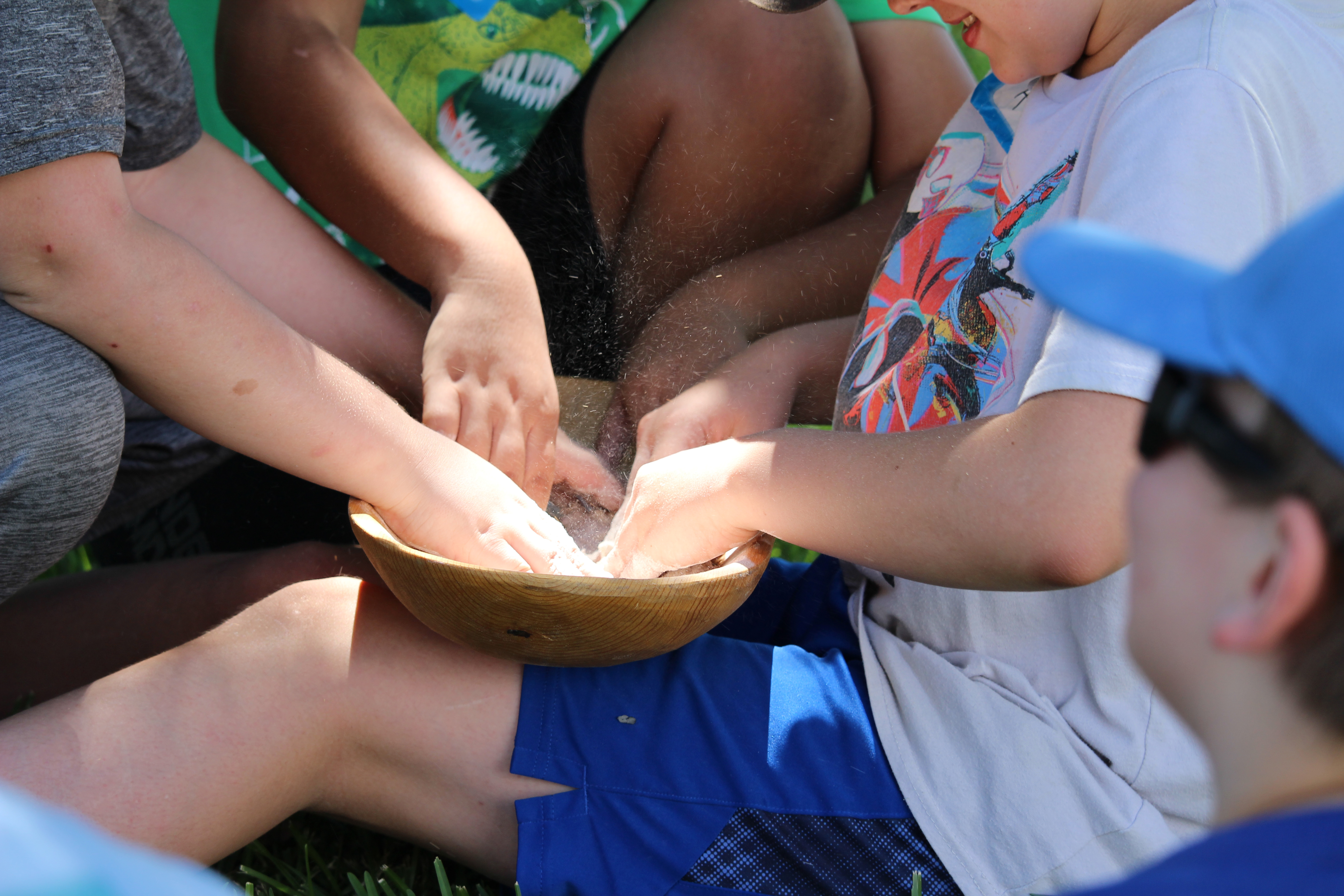 A group of kids holding a wooden bowl in one of their laps mix bread dough in the bowl.