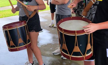 Two military drums are played by kids while other kids watch.