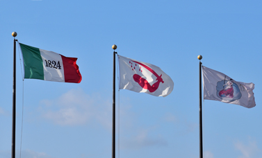 Three flags hang from flag poles: 1824 flag, Bloody Arm flag, San Jacinto flag.