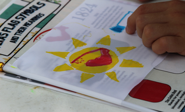 A child's hand colors a design with a yellow sun on a paper laid over an illustrated paper about flag symbols.