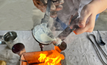 Hands pour a white liquid from a ladle into a mold, over a fire.