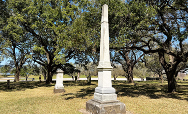 Photo of two tall stone cemetery markers near a grove of trees.