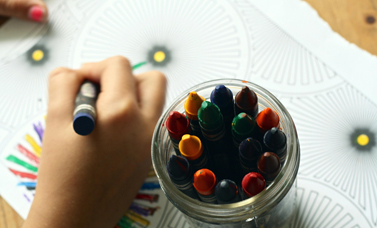 A hand holds a crayon over a coloring page, with a jar of crayons near.