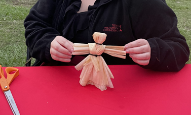 A person holds a corn husk doll upright, by its arms.