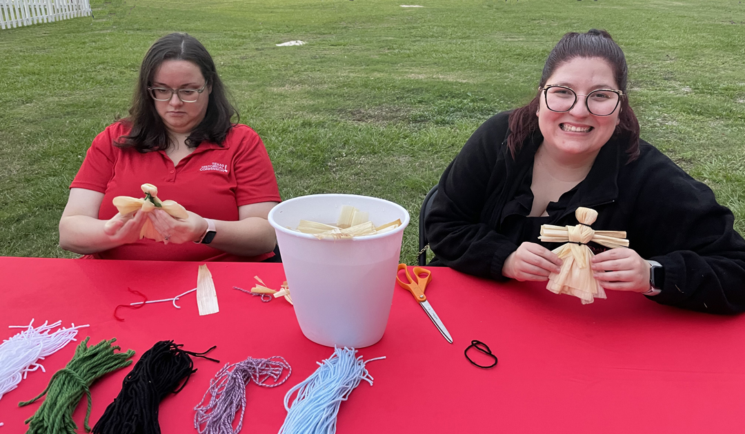 Two women sitting at a table hold corn husk dolls, with a pail of corn husks and piles of yarn on the table.