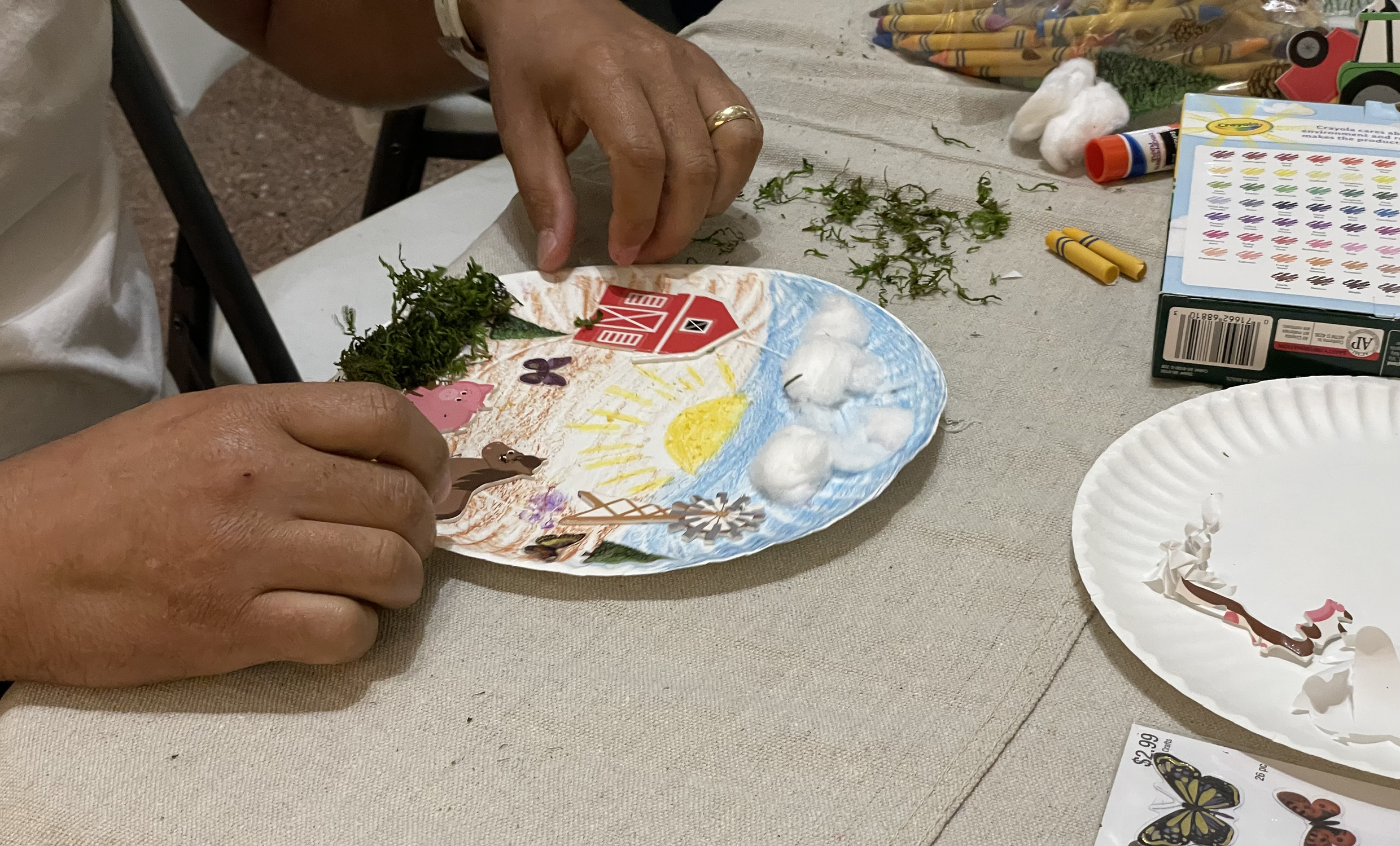 Hands create a three dimensional diorama of a barn and horse, on a paper plate.