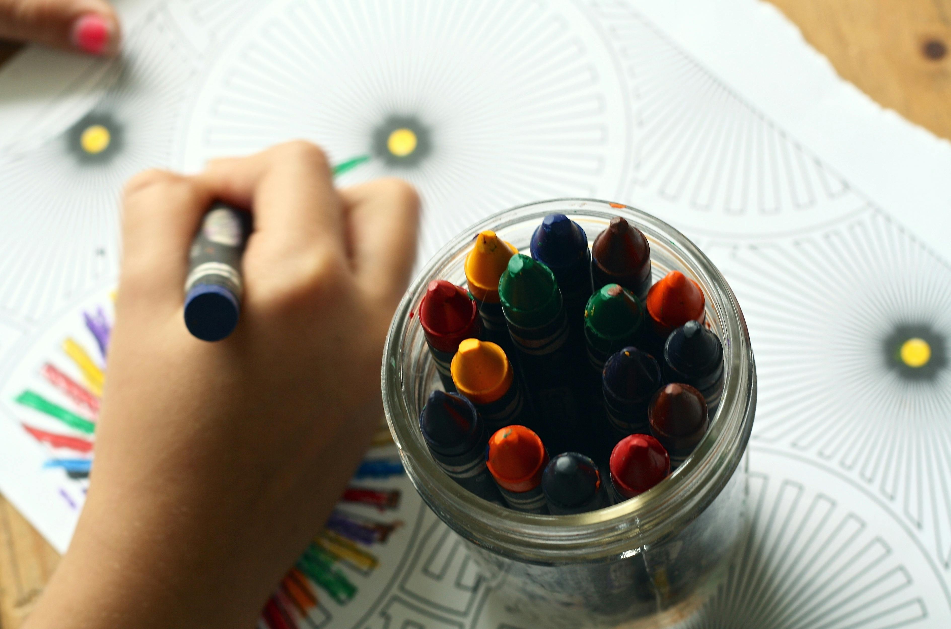 A hand holds a crayon over a coloring page, with a jar of crayons near